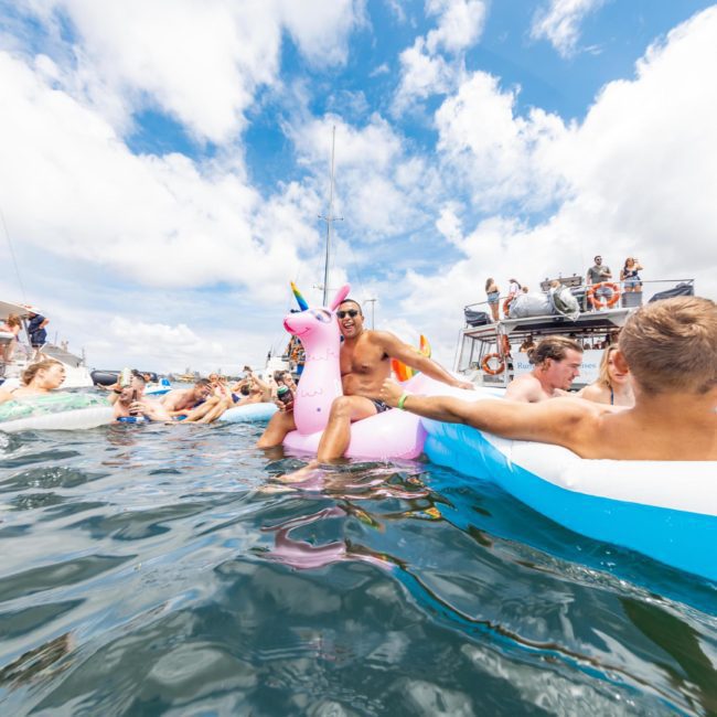 A group of people on floats, including a person on a pink unicorn float, enjoy a sunny day on the water near several boats for hire. Perfect for those considering DJ boat hire Sydney or looking to partake in a Catamaran party Sydney.