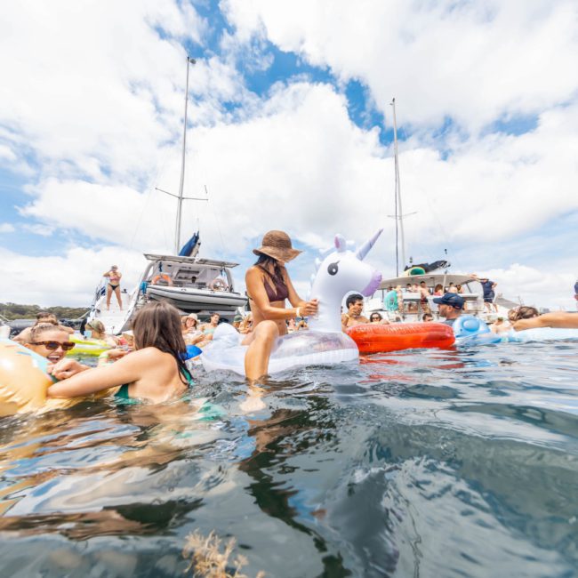 A group of people enjoys a day on the water, floating near boats and using inflatables, including a large inflatable unicorn. The sky is partly cloudy with a blue horizon in the background during their private yacht charter Sydney Harbour event.