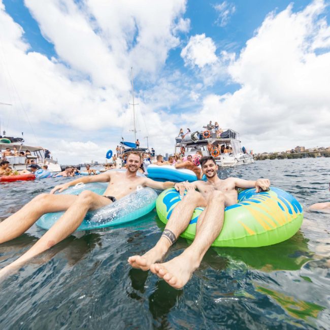 Three men in swimwear are floating on inflatable tubes in the water, surrounded by boats and people. The sky is partly cloudy, and everyone appears to be enjoying the water activities at a lively Sydney boat party hire.
