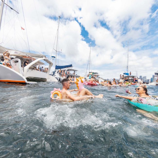 People floating in water with inflatable rings near boats on a sunny day, enjoying a vibrant DJ boat hire party in Sydney.