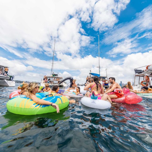 People relaxing on colorful inflatable rings in the ocean near several boats, including a private yacht charter in Sydney Harbour, on a sunny day with a partly cloudy sky.