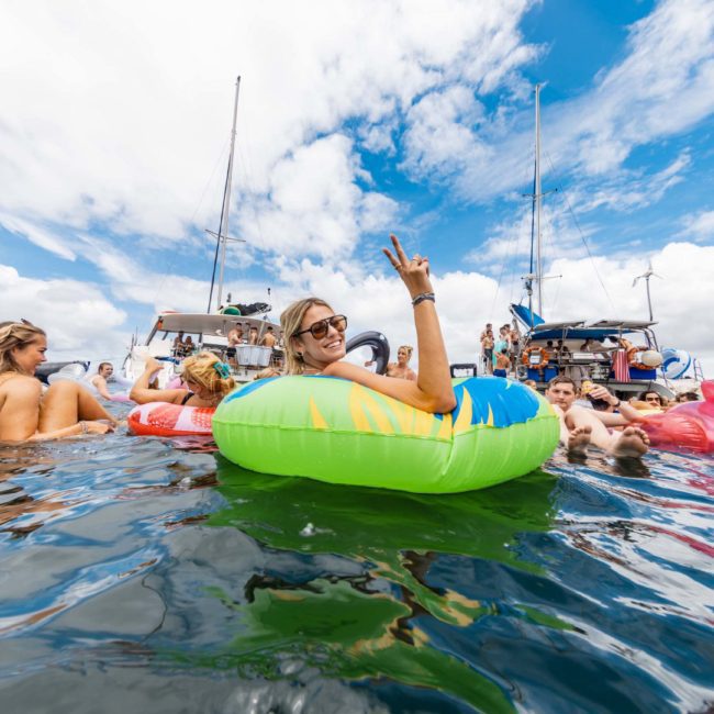 A group of people float on inflatable rings and relax in the water near anchored boats on a sunny day. One person in the foreground is smiling and flashing a peace sign during a luxury yacht hire Sydney event.