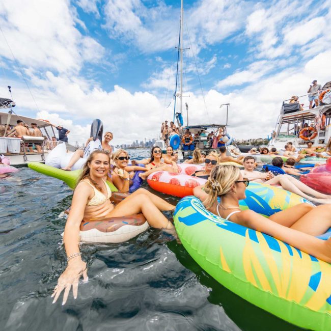 A group of people floats on colorful inflatable tubes in the water near docked boats, enjoying a partly cloudy sky. For an added touch of fun, consider a Sydney boat party hire to elevate your experience.