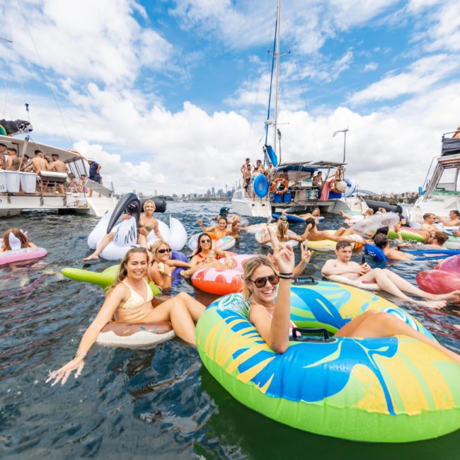 People in swimsuits are floating on inflatable rafts and pool tubes in the water near several boats on a sunny day, creating the perfect atmosphere for a Private yacht charter Sydney Harbour or even a DJ boat hire Sydney.