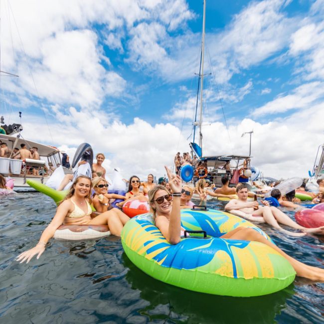 People relaxing and socializing on inflatable floats and boats in the water on a sunny day, with a backdrop of other anchored boats and a partly cloudy sky during a lively catamaran party in Sydney.