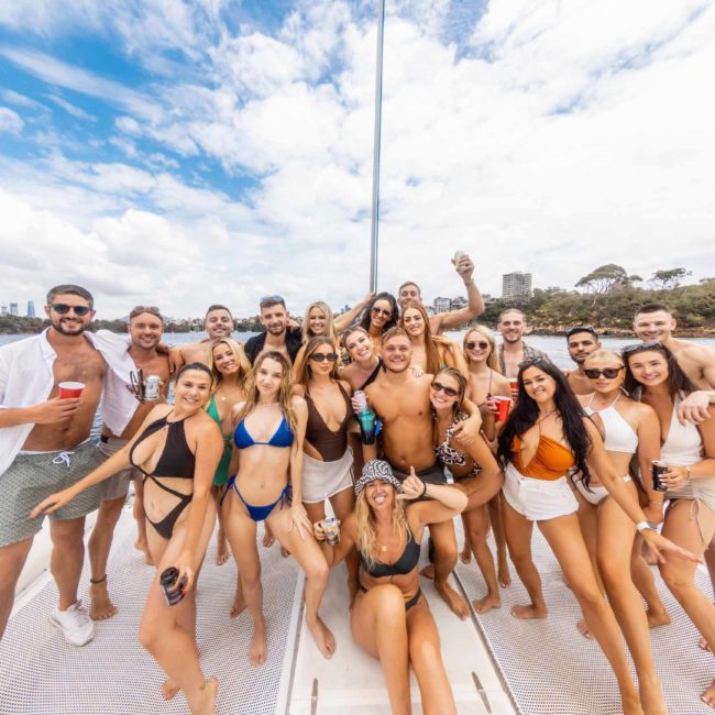 A large group of people in swimwear, posing on a boat with water and cityscape in the background on a sunny day, enjoying their Sydney boat party hire.