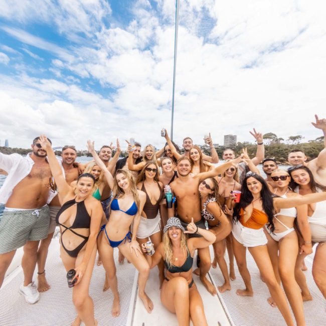A large group of people in swimwear are posing and smiling on a luxury yacht hire in Sydney. The background shows water, trees, and a city skyline under a partly cloudy sky.
