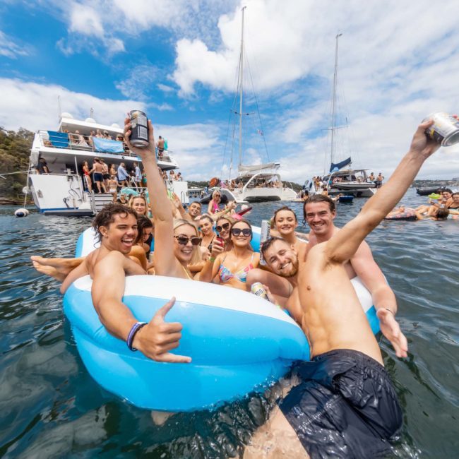 A group of people in swimsuits are enjoying themselves on an inflatable in the water next to boats, holding drinks. The sky is partly cloudy, and others are nearby in the water and on inflatables. It's a perfect scene for a Sydney boat party hire.