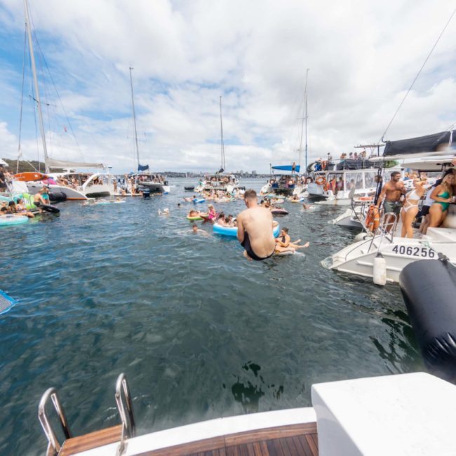 People and boats gathered in a bay, with individuals swimming and socializing on various floating platforms. One person is in mid-air, jumping into the water. Experience the ultimate Sydney boat party hire and enjoy unforgettable moments aboard a luxury yacht.