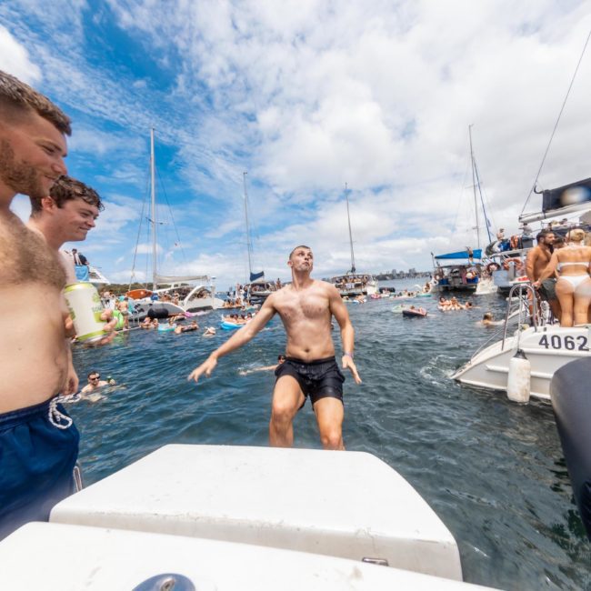 A shirtless man jumps into the water from a boat while other people, mostly in swimwear, socialize on boats and in the water under a sunny sky during a Sydney boat party hire.