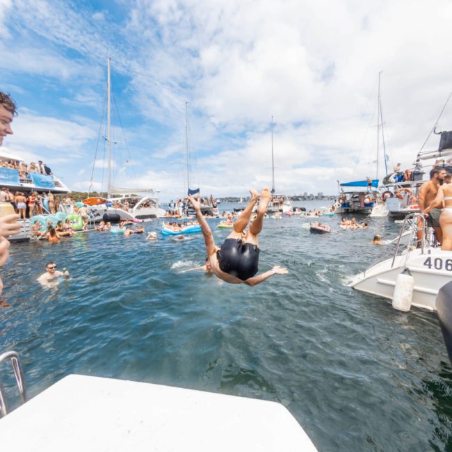 A man mid-air after jumping from a boat into a crowded lake area with people on various boats and in the water under a partly cloudy sky, capturing the vibrant atmosphere typical of a Sydney boat party hire event.