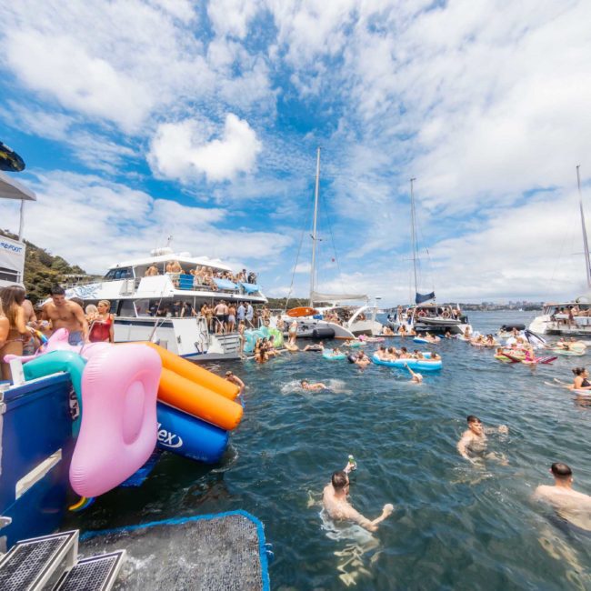 People are gathered in and around boats and inflatables on a sunny day, enjoying a lively catamaran party on Sydney Harbour.