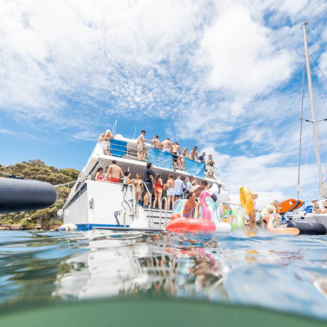 People are gathered on the deck of a boat, with more individuals swimming and floating on various inflatable toys in the water below. The partly cloudy sky adds to the fun ambiance of a Catamaran party Sydney event.