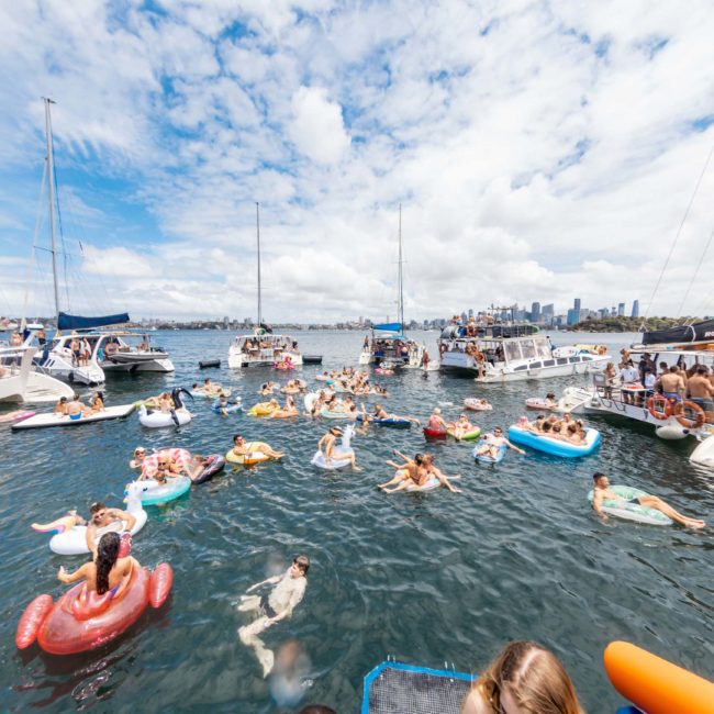 People in colorful inflatable rafts and boats gather on the water near multiple anchored motorboats and sailboats, enjoying a vibrant atmosphere against a backdrop of a city skyline under a partly cloudy sky, perfect for corporate boat events Sydney.