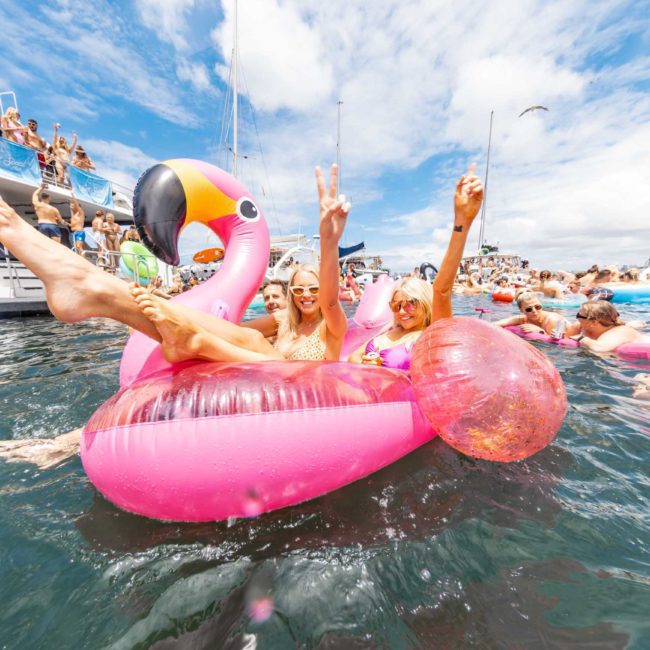 Three people enjoying themselves on a large pink flamingo float in the water, surrounded by other swimmers and boats under a partly cloudy sky, all part of a vibrant Sydney boat party hire.
