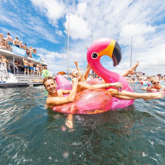 People are enjoying a sunny day on the water, with some swimming and others resting on a large inflatable pink flamingo, near a catamaran party in Sydney. The boat is crowded with more people reveling in the festivities.
