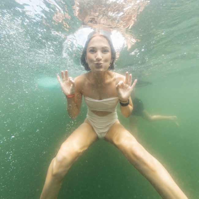 A person in a white swimsuit poses underwater with their hands raised and cheeks puffed out during a lively catamaran party in Sydney.