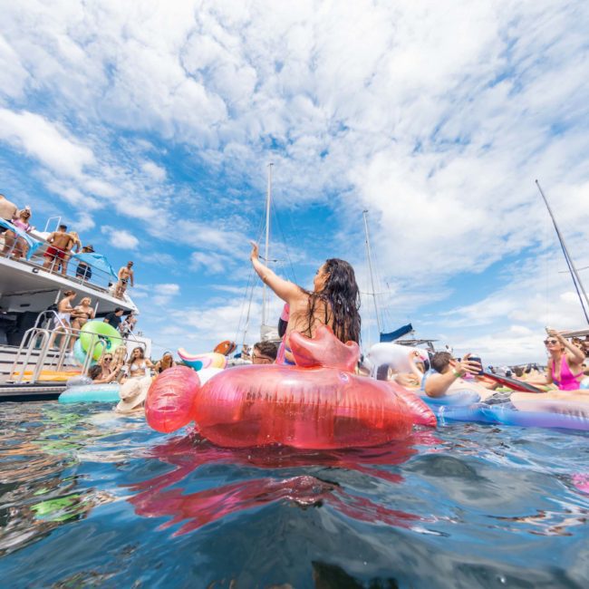 A group of people on inflatable pool floats enjoying a sunny day on the water, with several boats docked nearby and a partly cloudy sky overhead, perfect for those considering DJ boat hire Sydney or private yacht charter Sydney Harbour.