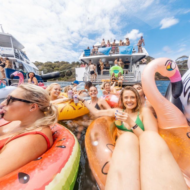 A group of people enjoying a sunny day on inflatable pool floats with beverages, anchored near large boats. The scene is lively with smiling faces and colorful inflatables, including a flamingo and watermelon. It’s the perfect setting for a Catamaran party Sydney or private yacht charter Sydney Harbour.