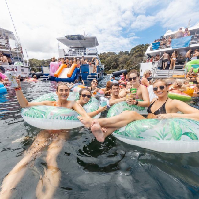 People are in an outdoor water setting, floating on inflatable rafts, holding drinks, and surrounded by boats with others. The environment is lively and social under a partly cloudy sky, perfect for a luxury yacht hire Sydney experience or corporate boat events Sydney.