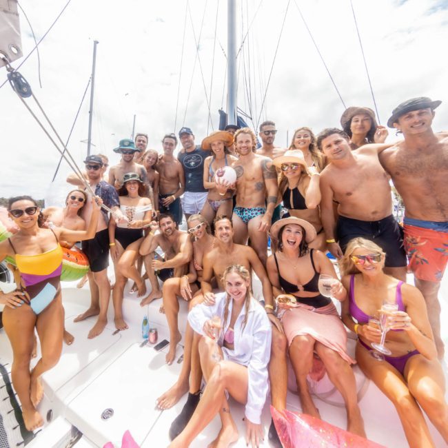 A large group of people in swimwear gather on a catamaran party in Sydney, enjoying a sunny day. Most are smiling and holding drinks, with a cityscape visible in the background.
