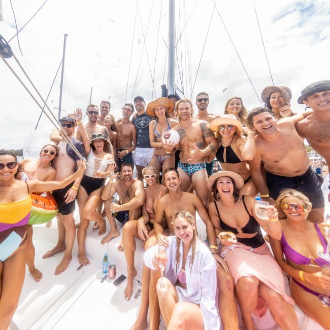 A large group of people in swimwear smiles and poses together on a private yacht charter Sydney Harbour under a bright sky.
