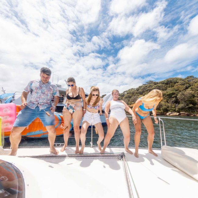 A group of five people in swimwear stand on a boat deck near the water, with inflatable items and blue sky visible. Two more people, also in swimwear, are sitting on the side of the deck. Enjoy a corporate boat event or luxury yacht hire in Sydney Harbour for a memorable experience.