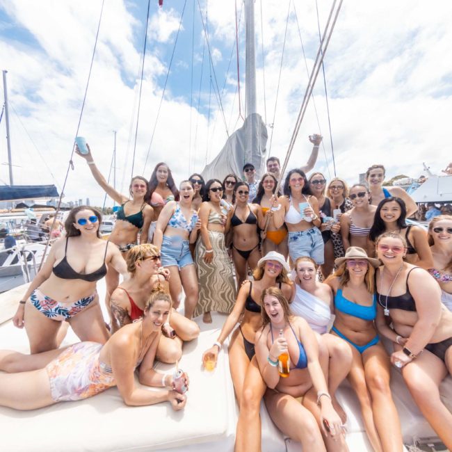 A group of people in swimwear poses for a photo on a boat deck under a partly cloudy sky. Some are standing, others sitting or lying down, with boats visible in the background, enjoying a private yacht charter Sydney Harbour experience.