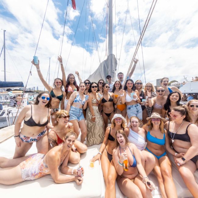 A large group of people in swimwear pose together on a boat docked at a marina, with sailboats and clear skies in the background, enjoying a private yacht charter Sydney Harbour.