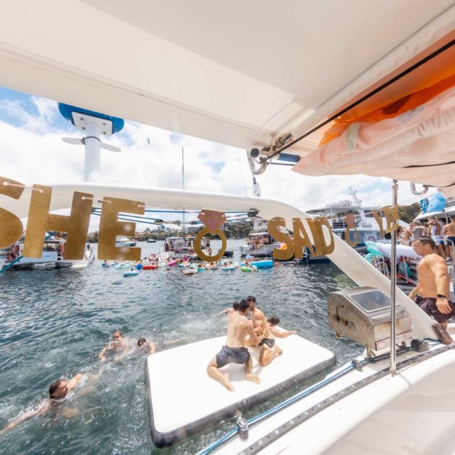 People enjoying a day on a catamaran party in Sydney Harbour with a "She said yes" banner; some people are swimming while others relax on a floating platform in the water.