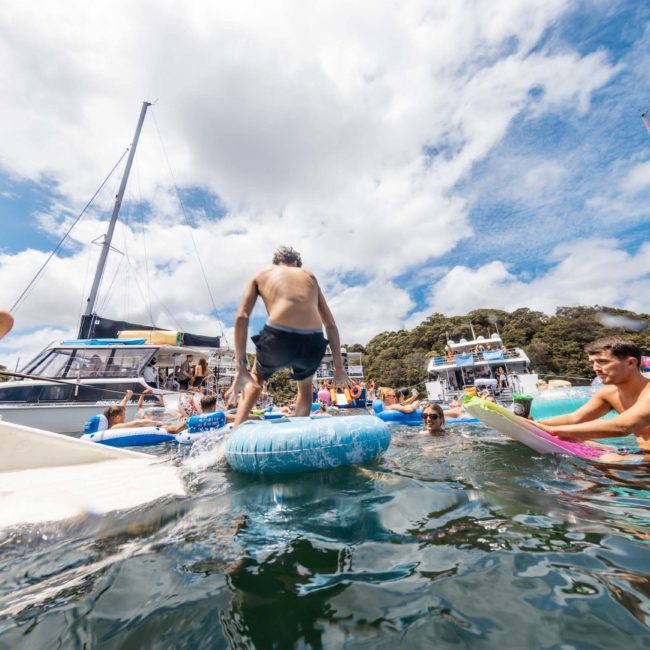 People enjoying a sunny day on the water with boats and inflatables; a person jumping into the water from an inflatable tube while others look on during a corporate boat event in Sydney.