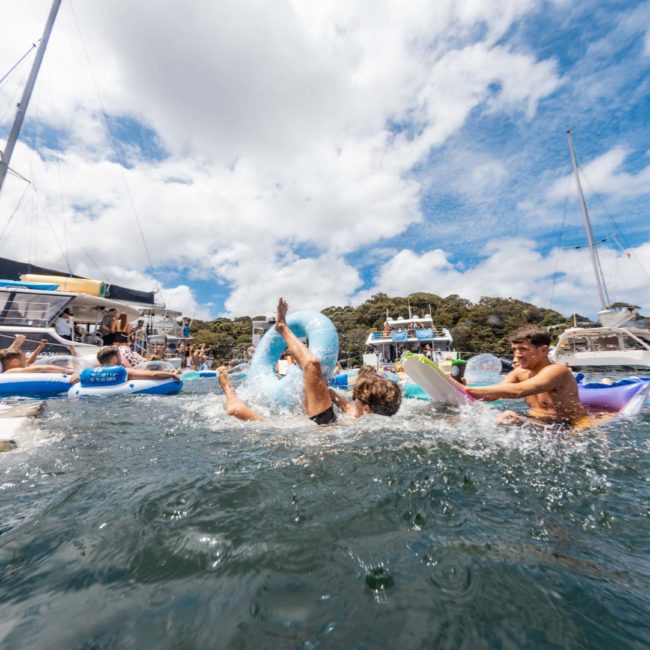People on inflatable floaties play in the water with several yachts and boats anchored nearby under a partly cloudy sky, creating the perfect setting for a luxury yacht hire Sydney experience.