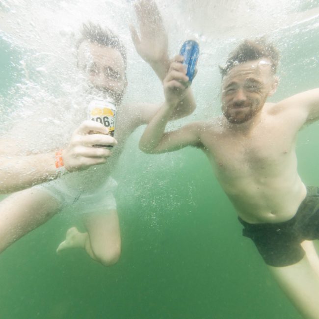Two men underwater holding cans, surrounded by bubbles. One man in white shorts and the other in black shorts, both smiling and extending their free hands toward the camera during a lively Sydney boat party hire.