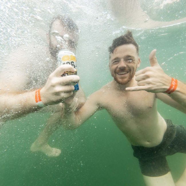 Two shirtless men underwater, both wearing red wristbands. One man is holding a beverage can, while the other is making a "hang loose" hand gesture. Perfect snapshot from a Catamaran party Sydney style!
