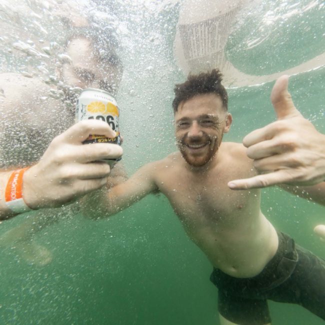 Two men underwater, one holding a can of beer and making a hand gesture with his other hand, both wearing swim trunks and wristbands. Luxury yacht hire Sydney makes for an unforgettable setting for this aquatic fun.