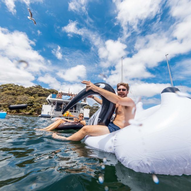 Two people enjoy a sunny day by the water, lounging on a large inflatable float. A seagull flies overhead, and boats, including luxury yacht hires in Sydney, are anchored in the background.