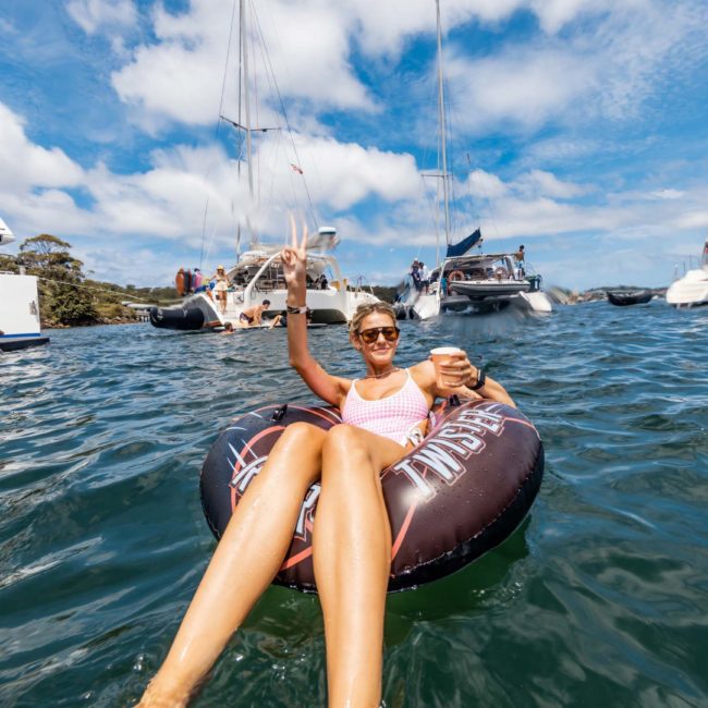 A person relaxes in an inflatable tube holding a drink on a sunny day, surrounded by boats and water, while nearby, others enjoy the elegance of a private yacht charter on Sydney Harbour.