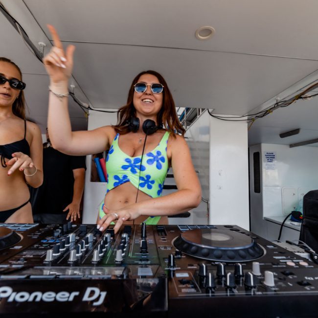 Two women in swimwear stand behind a DJ booth on a luxury yacht hire in Sydney, with one woman pointing into the air and the other adjusting controls on the DJ equipment.