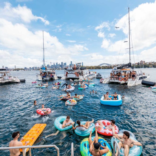 A group of people enjoy a sunny day on the water, lounging on inflatables and swimming near several docked boats. The city skyline is visible in the background, with a luxurious yacht hire Sydney adding to the perfect atmosphere.