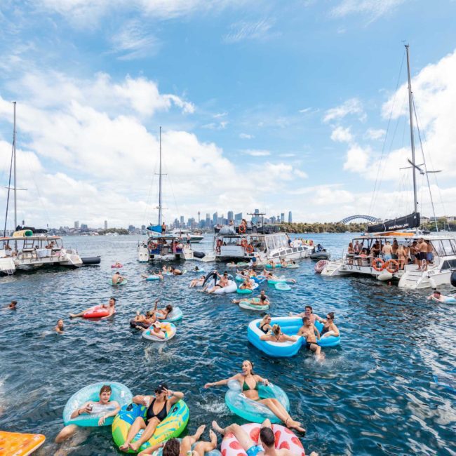 People are enjoying a sunny day on the water, floating on inflatables and swimming near anchored boats with a city skyline in the background, while a luxurious private yacht charter cruises through Sydney Harbour.