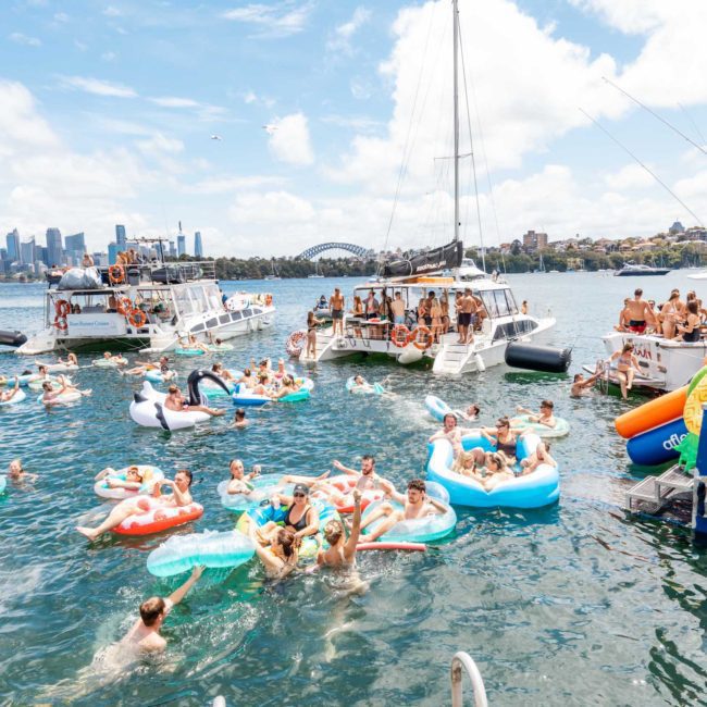 A vibrant scene of people on boats and inflatables enjoying a sunny day on the water with a cityscape in the background, perfect for a Sydney boat party hire or private yacht charter Sydney Harbour.