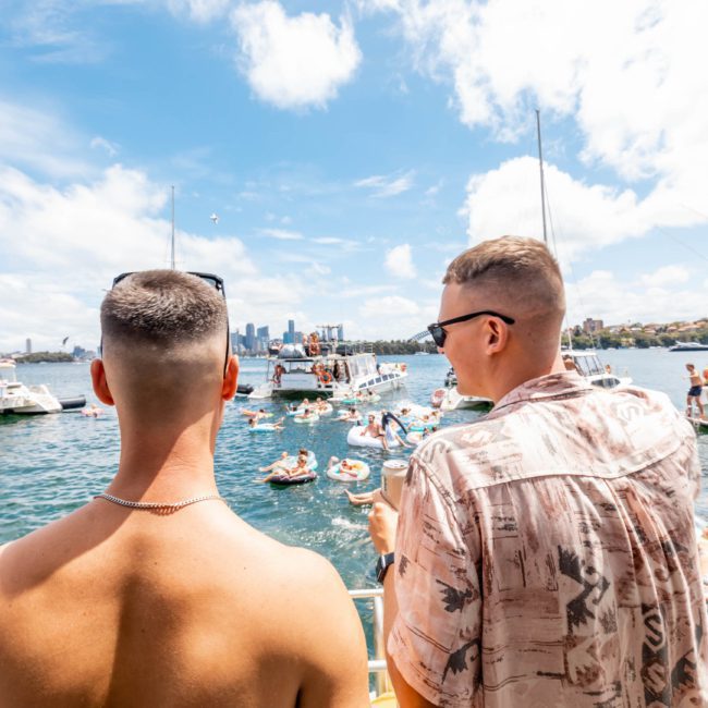 Two men on a private yacht charter in Sydney Harbour observe a crowd of people on floaties and other boats in the sunny, bustling harbor.