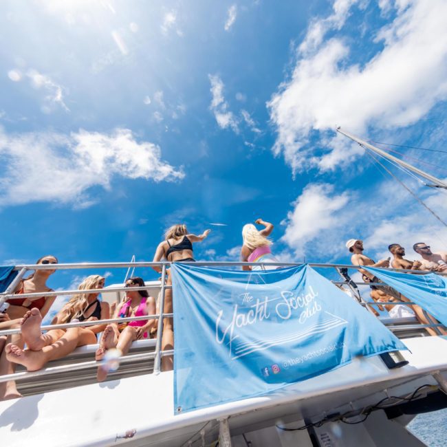 People are on a yacht with "The Yacht Social Club" banners, under a blue sky with scattered clouds. Some are sitting and some are standing, enjoying the view and the sunny weather. Perfect for a corporate boat event on Sydney Harbour or those seeking luxury yacht hire in Sydney.