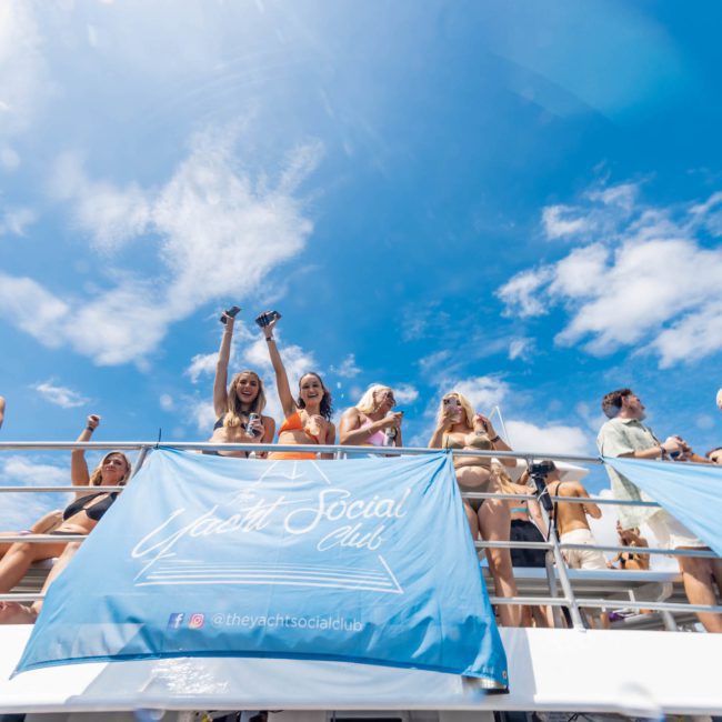 A group of people enjoying a sunny day on the upper deck of a boat, with banners stating "The Yacht Social Club" hanging along the railing. White clouds and blue sky form the background, perfect for a Sydney boat party hire.