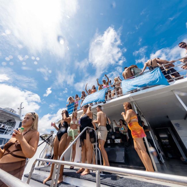 A group of people in swimwear enjoys a sunny day on the deck of a private yacht charter in Sydney Harbour, with some of them dancing and others relaxing. The sky is clear with a few clouds.