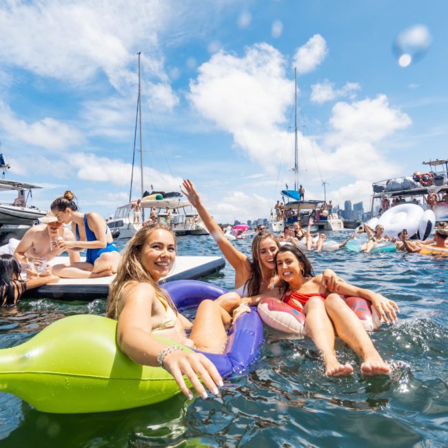 Three women floating on inflatable rafts in the water, surrounded by other people and boats under a partly cloudy sky, enjoying a luxury yacht hire Sydney experience.