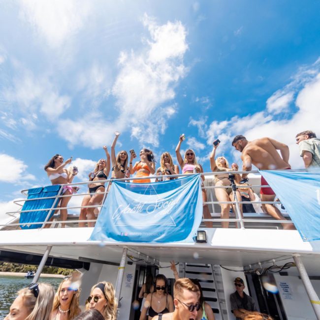 A group of people on a boat deck under a blue sky, some dancing and others engaging in conversation. Blue banners are displayed on the railings, perfect for a catamaran party in Sydney.