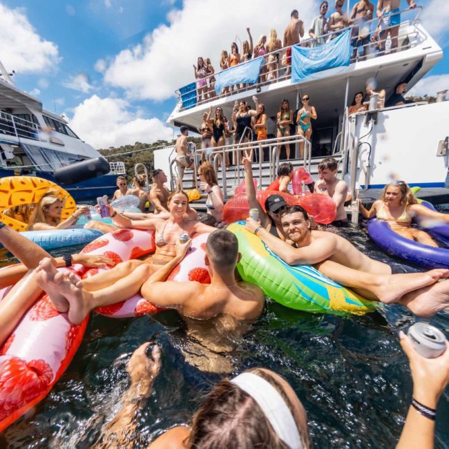 People enjoy a sunny day on a boat, with many of them floating in the water on colorful inflatables, while others are on the deck. The background shows clear skies and calm waters. It’s the perfect scene for a luxury yacht hire in Sydney.