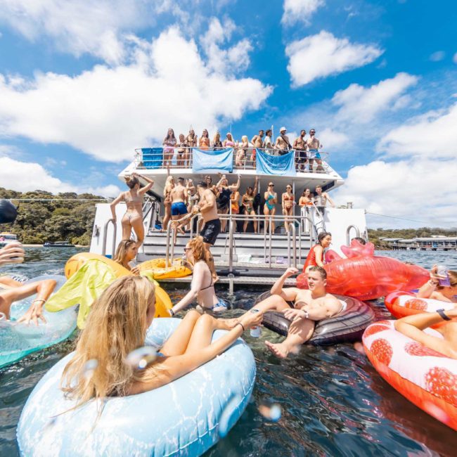 A lively group of people on and around a boat, many floating on inflatable rafts in the water, under a partly cloudy sky. Experience the best of a Sydney boat party hire or enjoy the luxury of a private yacht charter on Sydney Harbour.