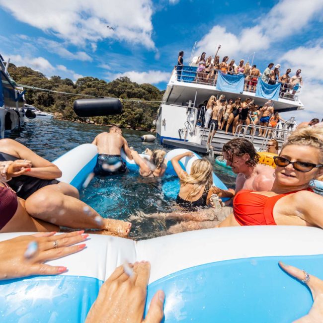 People enjoying a sunny day on a large inflatable raft in the water, with a lively boat event in the background. Some are swimming, while others relax or socialize. Trees and blue sky are visible.